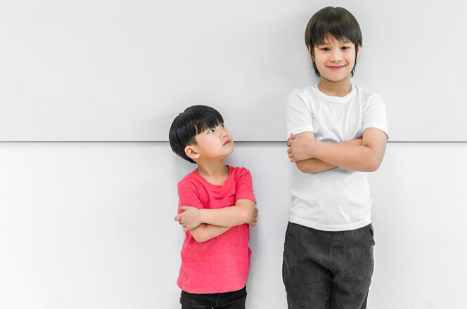 
					Little child boy standing arms crossed and looking face of tall child at standing arms crossed and smiling. Big and small kid concept at be friends.